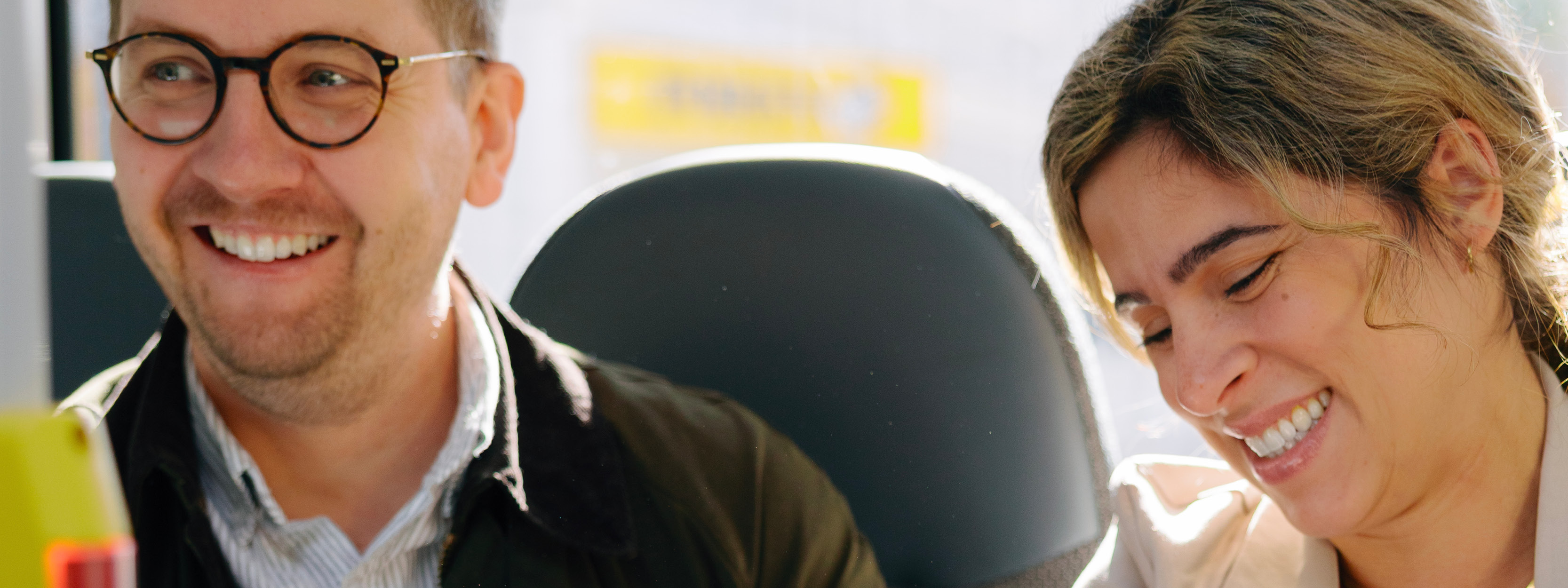 En man och en kvinna sitter på en buss. Mannen sitter till vänster. Kvinnan till höger. De ler. 