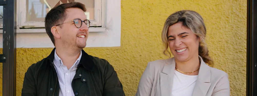 En man och en kvinna sitter på en busshållplats. Mannen ler. Kvinnan ler. 