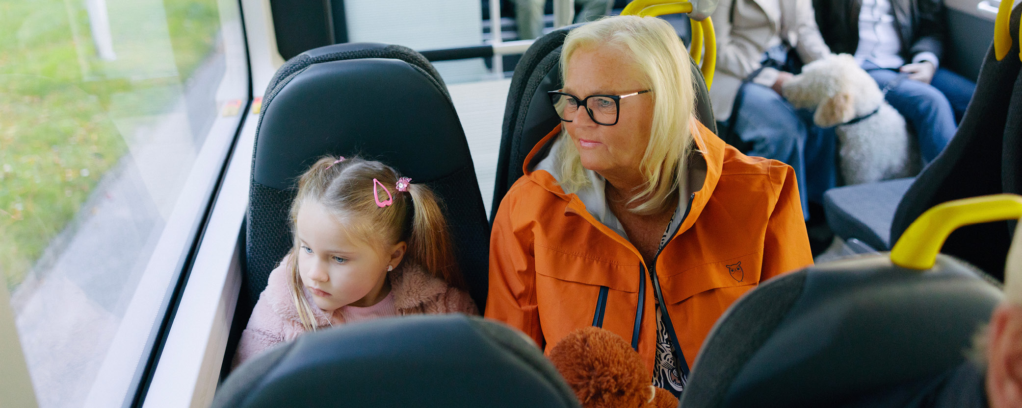 En liten tjej sitter utmed fönstret i en buss. Bredvid henne sitter en dam i orange jacka.