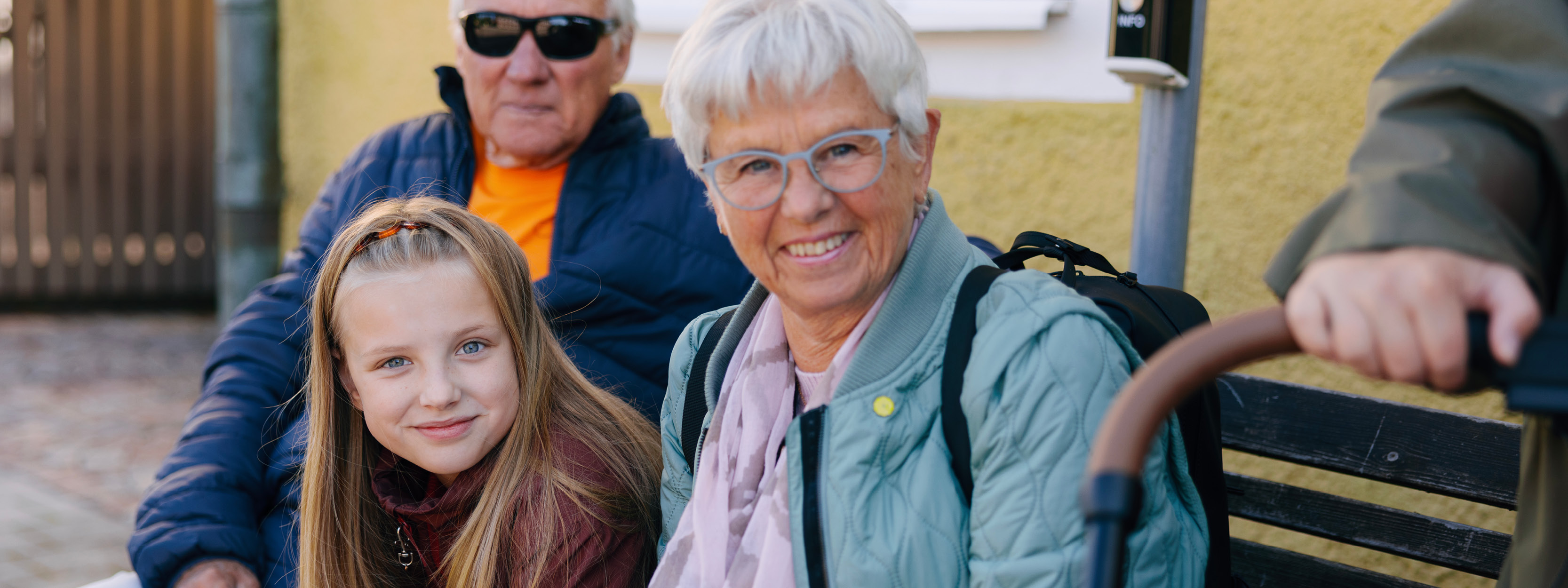 En dam sitter och ler. Bredvid henne sitter en tjej. I bakgrunden syns en man med solglasögon.