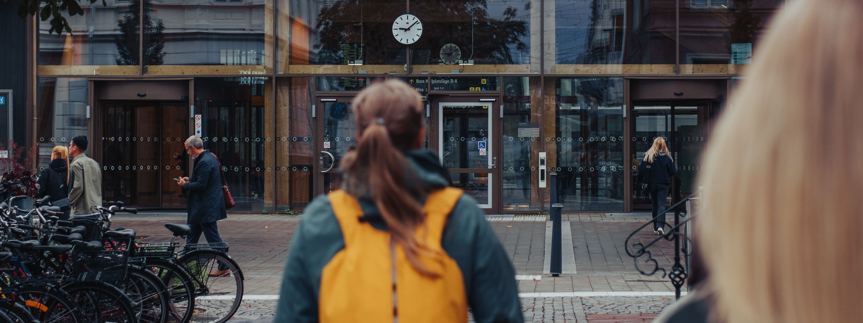 En kvinna går. Hon bär en gul ryggsäck. I bakgrunden syns en station och människor i rörelse. 