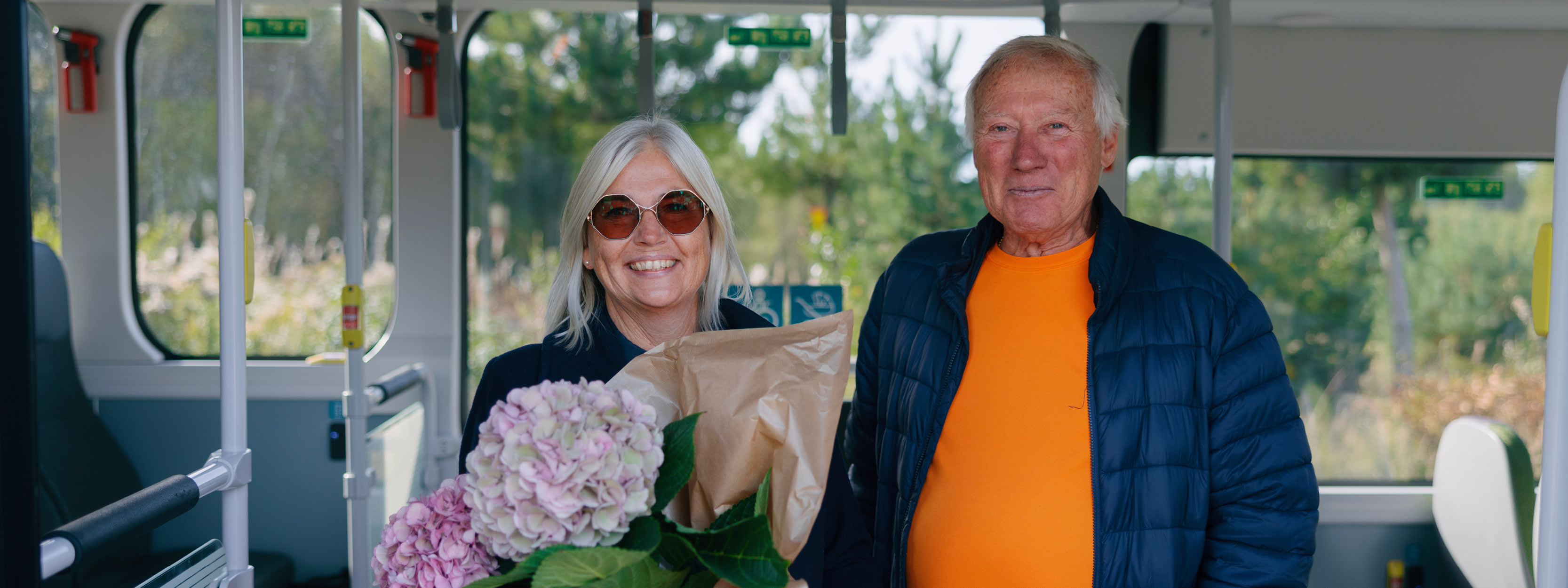 En man och en kvinna står i dörröppningen på en buss. De ler. Kvinnan bär på blommor. 