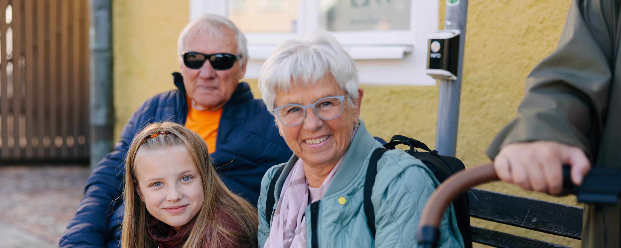 En dam sitter och ler. Bredvid henne sitter en tjej. I bakgrunden syns en man med solglasögon. 