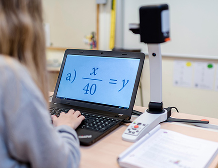 Ein Mädchen in einem Klassenzimmer verwendet die Lesekamera MagniLink S zusammen mit einem Computer und MagniLink PCViewer, um die Zahl in einem Mathematikbuch zu vergrößern.