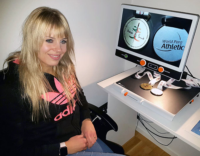 Nathalie sitter framför sin läskamera MagniLink Zip. På läsbordet ligger två medaljer som syns förstorade på Zip:ens skärm.