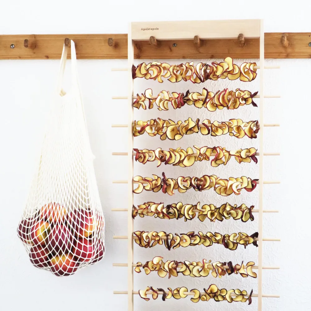 Torkade äppelringar hänger på Apelstapeln Äppeltork, bredvid hänger en nätpåse med röda äpplen