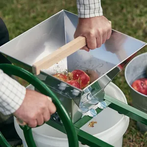 Vuxen man använder vevhjulet på en manuell fruktkross för att krossa röda äpplen utomhus i trädgården