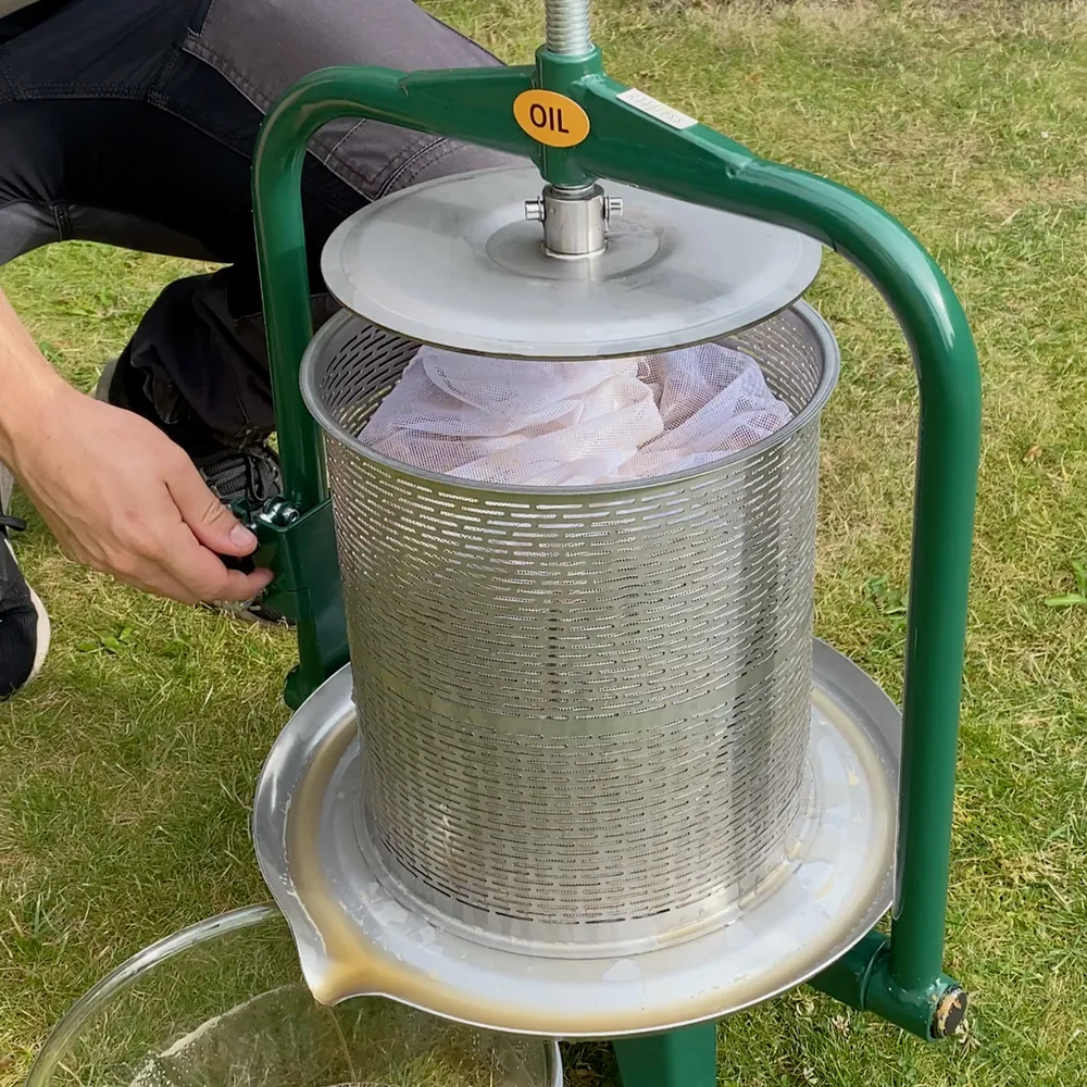 En vuxen mans hand läggs på silduken i en cylinderformad fruktpress som står utomhus