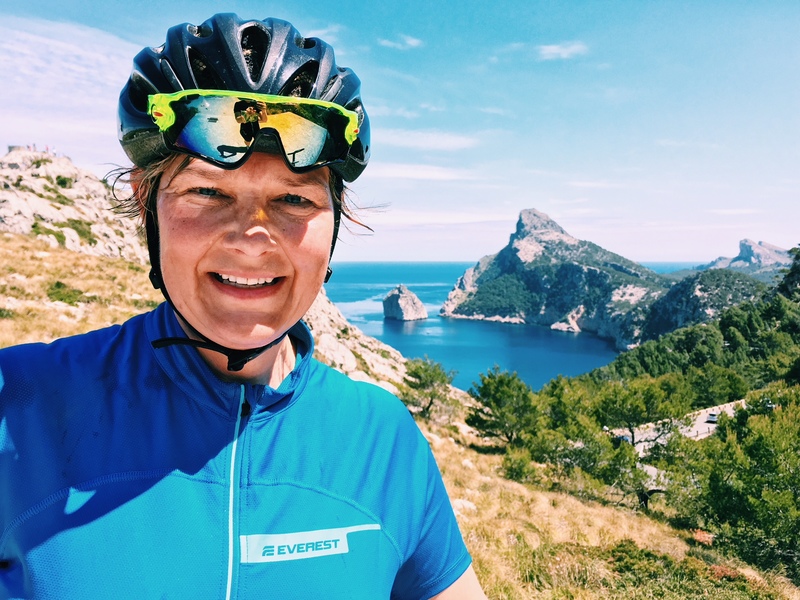 Porträttbild tagen i brösthöjd. Kvinna i cykelhjälm med hav och berg i bakgrunden. 