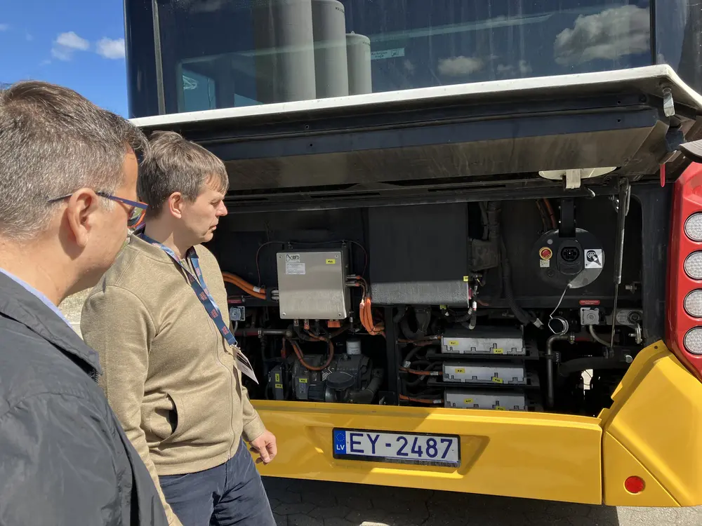 Två män står bredvid det öppna bakre utrymmet på en gul buss och undersöker dess exponerade motor och interna komponenter.