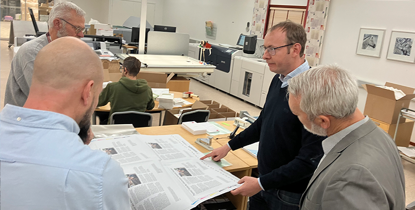 En man visar på en tryckt tidning i ett tryckeri, tre andra män tittar på.