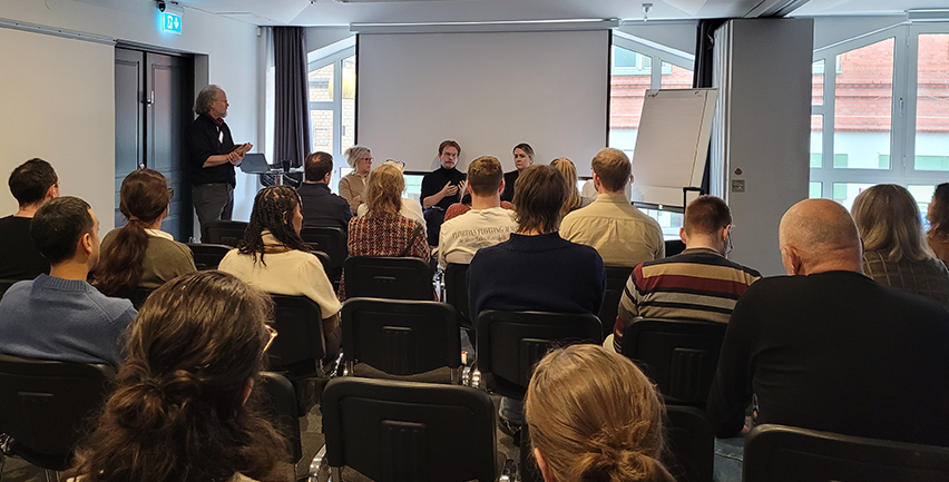 Människor sitter på stolar och lyssnar på fyra personer framför en vit duk.