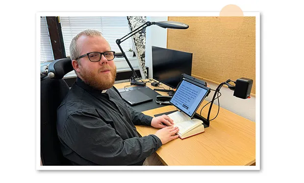 Linus Forsberg sitter på sitt kontor och använder läskameran MagniLink iTAB för att läsa texten i en bok.