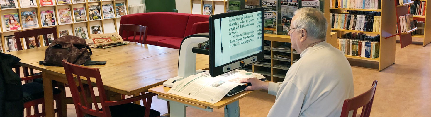 En äldre man på ett bibliotek sitter framför läskameran MagniLink Vision och läser tidningen.