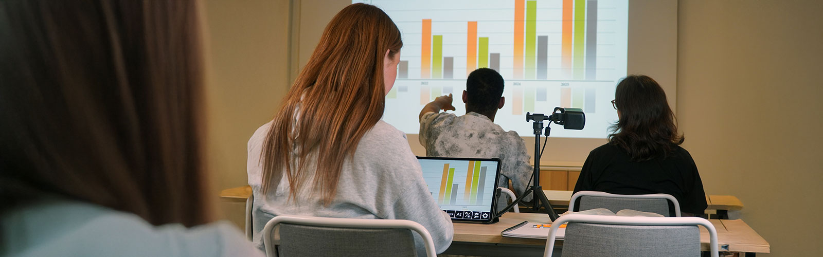 Backs of students are seen sitting in a classroom looking at a diagram on the board. A girl has a MagniLink Mini to see the diagram magnified on her computer screen.