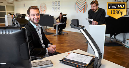 Billede af en smilende mand i et kontormiljø, der bruger LVIs læsekamera MagniLink Pro i sit arbejde ved skrivebordet.